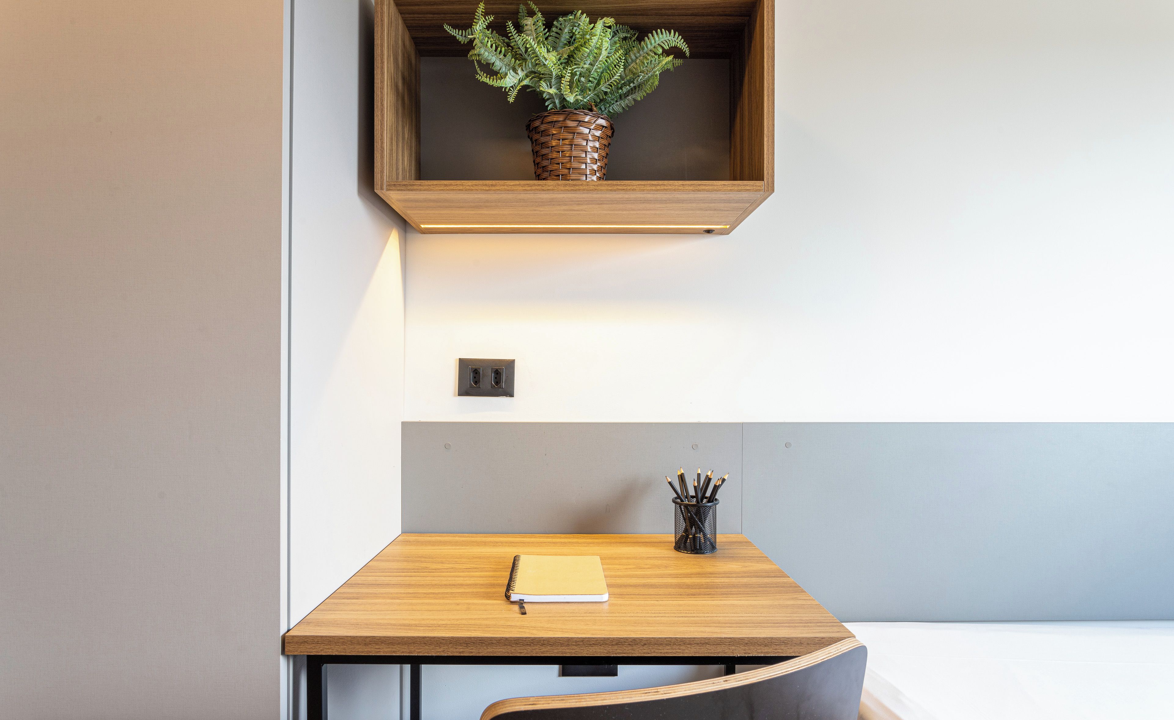 A small wooden desk with a notebook, a pen holder, and a potted plant on a shelf above.