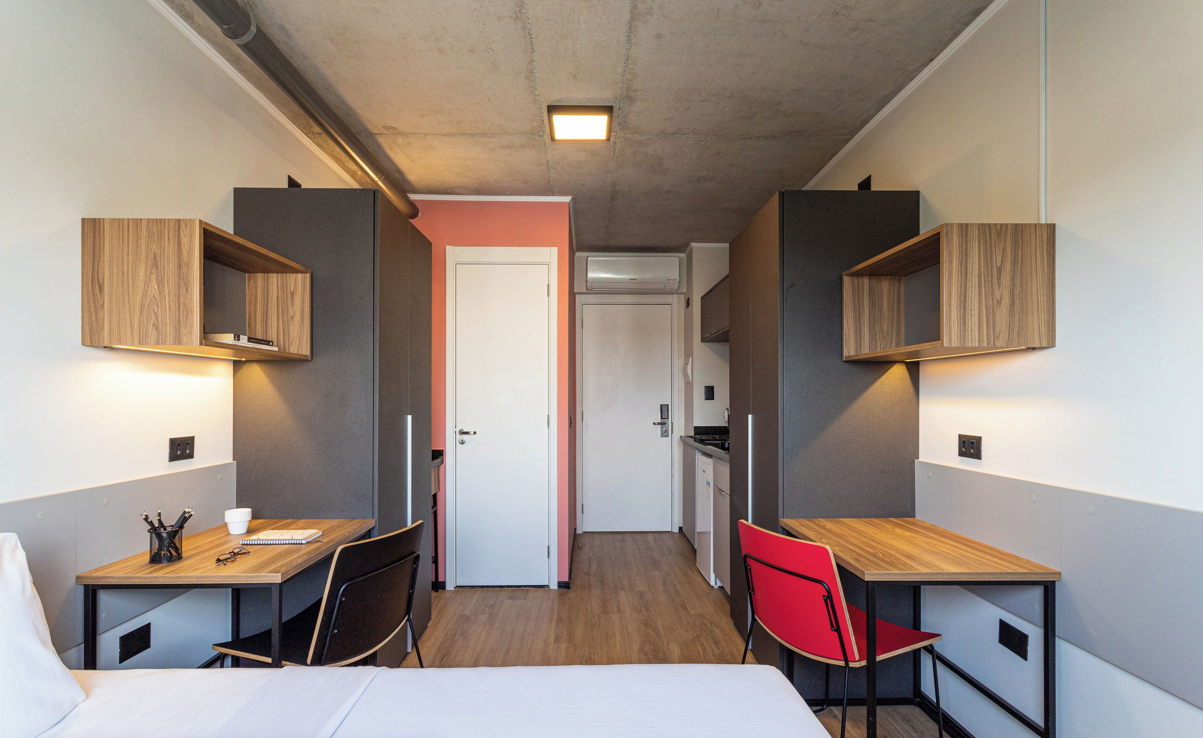 A compact hotel room with a bed, two desks with chairs, and built-in shelves.
