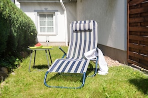 Ein gestreifter Liegestuhl mit einem Handtuch, ein kleiner Tisch mit einem Glas und einem Stück Wassermelone, sowie ein Hecke in einem Garten.