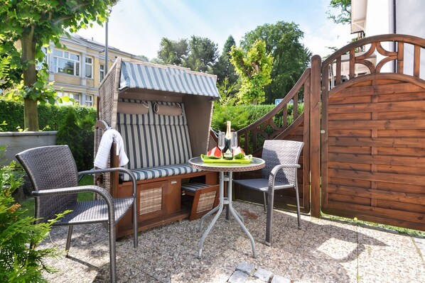An outdoor seating area with a covered bench, two chairs, and a table with a tray of drinks and watermelon.
