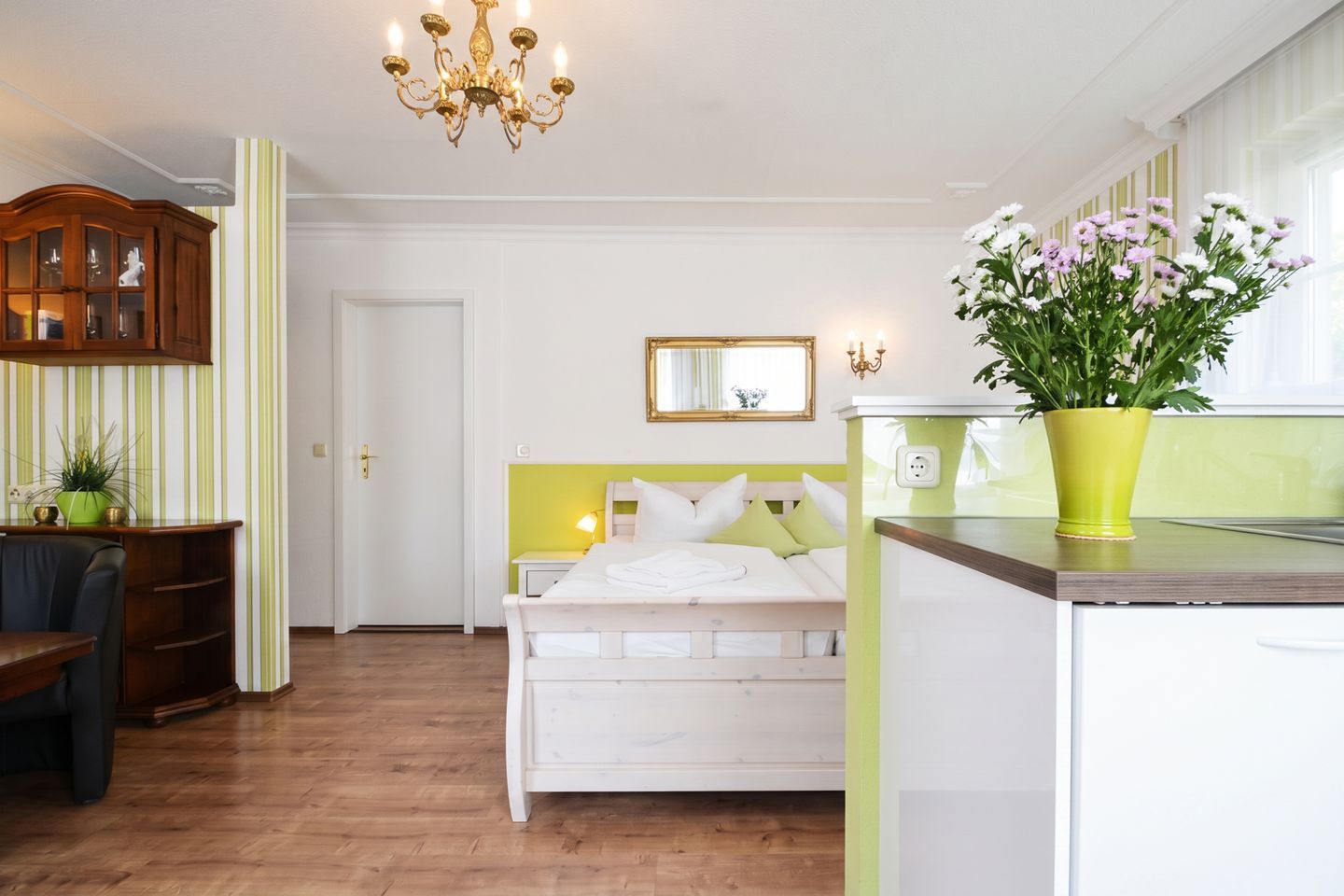 Une chambre d’hôtel avec un lit, un bureau, une chaise, un lustre, un miroir et une plante en pot.