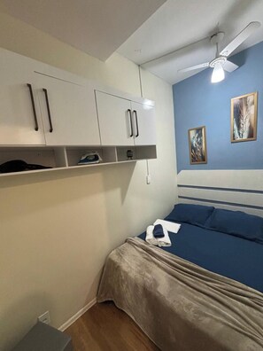 A bedroom with a bed, a ceiling fan, a shelf with items, and artwork on the wall. - Apartamento na Praia do Forte 😉 , Centro Cabo Frio - RJ 118 (Centro Cabo Frio)