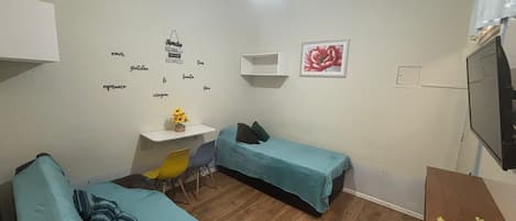 A small, well-lit room with two single beds, a desk with a chair, and a television.
