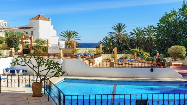 A pool area with a clear view of the sea, surrounded by palm trees and a white building with a tiled roof. - Fuengirola Seaside Bliss: 3 BR with a View (Fuengirola)