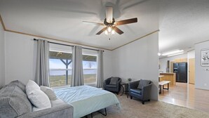 A bedroom with a bed, two armchairs, a small table, and a ceiling fan. There is a large window with curtains, a view of the outdoors, and a kitchenette in the adjacent room. - Big Parking Lot, Pets Invited, Fenced Farm Home (Kennewick)