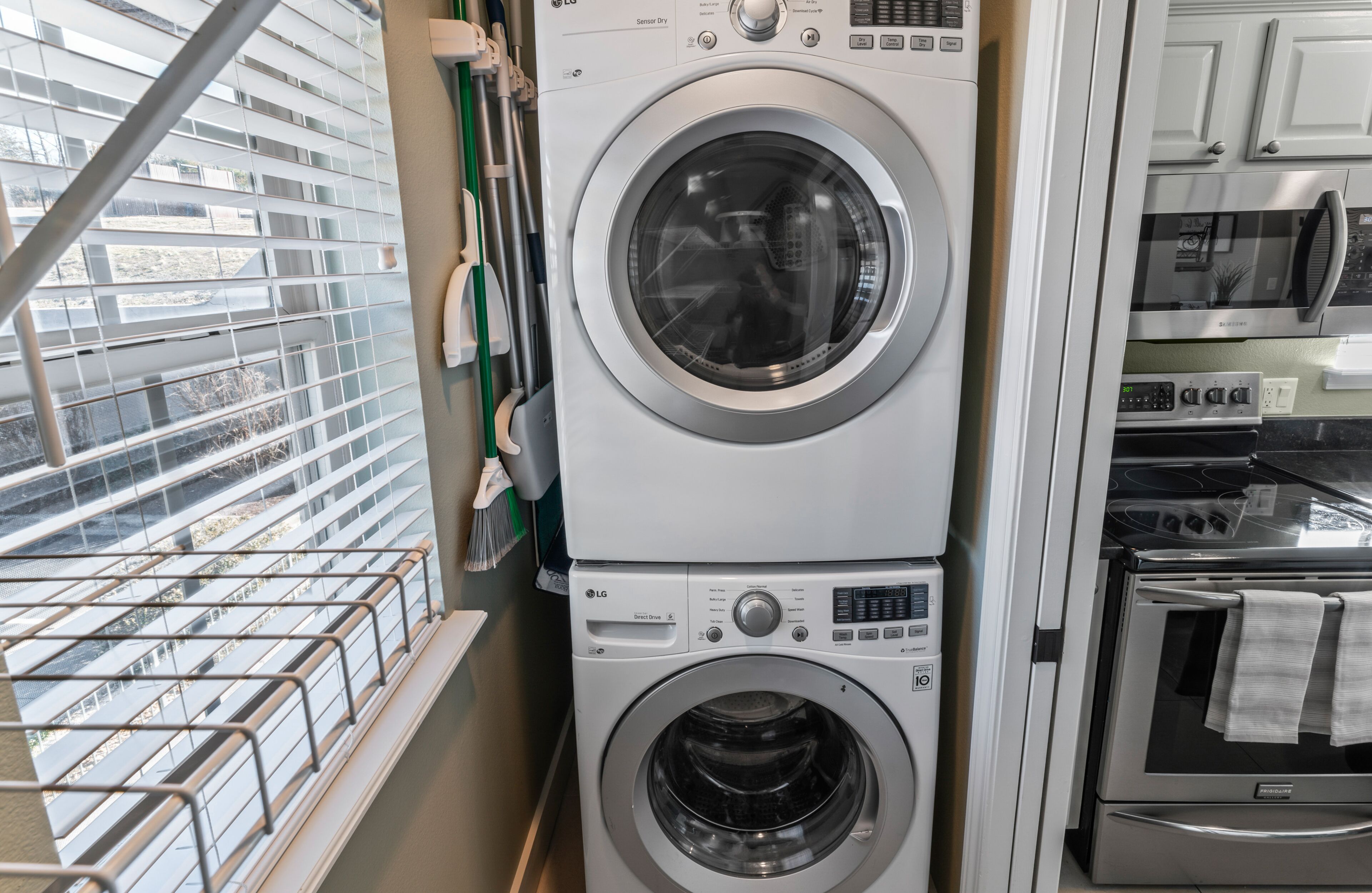 A laundry room with stacked washing machines and dryers, a window with blinds, and a view of a parking lot.