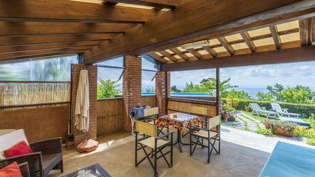 Eine überdachte Terrasse mit Tisch und Stühlen, ein aufgehängtes Handtuch und Blick auf Grünflächen und das Meer.
