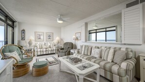 A living room with a white ceiling, a large window with shutters, a wicker chair, a striped sofa, and a coffee table. - Sea Terrace 88th St, Outdoor Pool, Oceanfront building & View, *FREE Linen Pkg* (Ocean City)