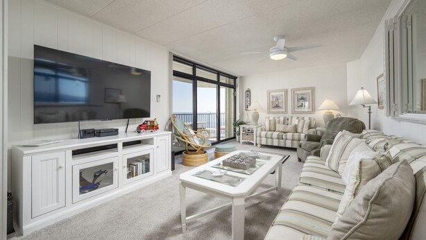 A spacious living room with a large flat-screen TV, a ceiling fan, and a balcony with a view. - Sea Terrace 88th St, Outdoor Pool, Oceanfront building & View, *FREE Linen Pkg* (Ocean City)