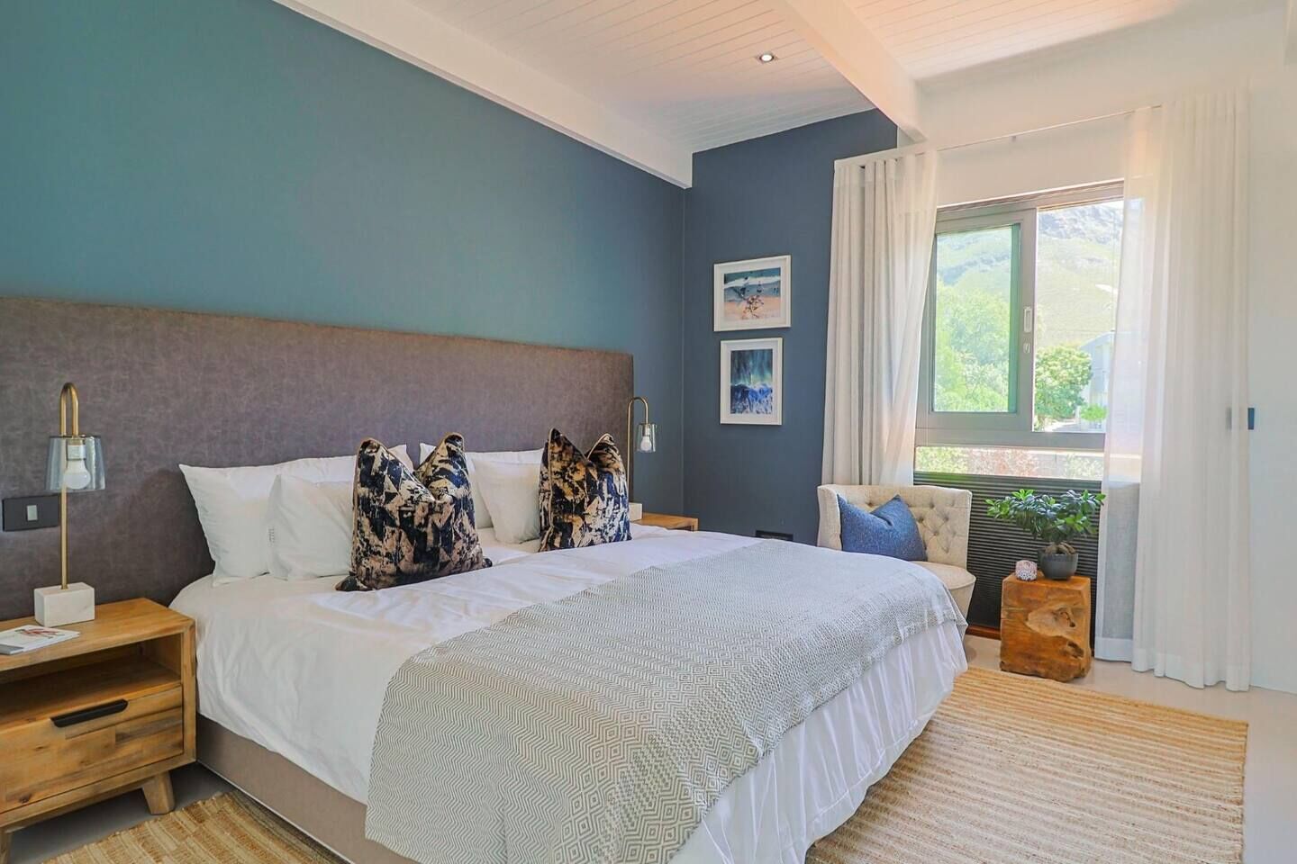 Une chambre avec un grand lit, des tables de chevet, une commode, une fenêtre avec des rideaux et une vue sur la verdure à l’extérieur.