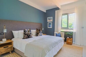 Une chambre avec un grand lit, des tables de chevet, une commode, une fenêtre avec des rideaux et une vue sur la verdure à l’extérieur.