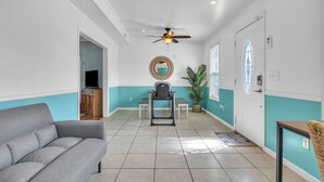 A spacious living area with a grey sofa, a ceiling fan, a small table with a chair, and a potted plant.
