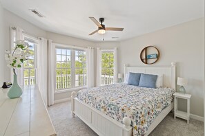 Une chambre à coucher comprenant un lit, un ventilateur de plafond, une fenêtre avec des rideaux, une table de chevet avec une lampe et un élément de décoration murale.