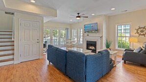 A spacious living room with a fireplace, a ceiling fan, and a large window. - "Pearl on a Strand" 6Bd/4Ba w/ Pool, Opt Golf Cart (Oak Island)