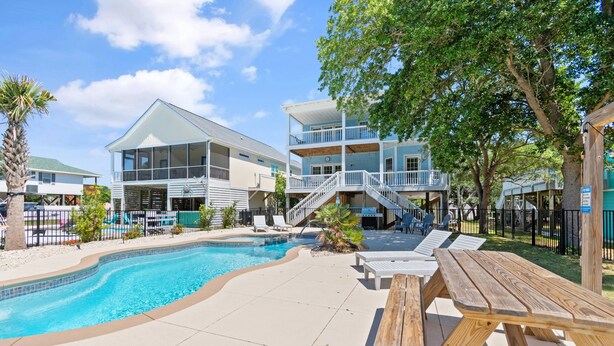 A pool area with a wooden deck, lounge chairs, and a multi-story building with balconies. - "Pearl on a Strand" 6Bd/4Ba w/ Pool, Opt Golf Cart (Oak Island)