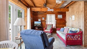 A log cabin interior with a blue armchair, a red wicker sofa, a television, and a 'WELCOME' sign.