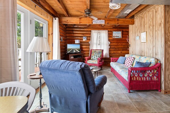 A log cabin interior with a blue armchair, a red wicker sofa, a television, and a 'WELCOME' sign.
