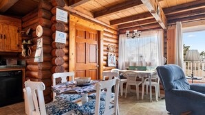 A cozy cabin interior with a wooden log wall, a dining area with a table and chairs, a kitchen with a refrigerator, and a large window offering a view.