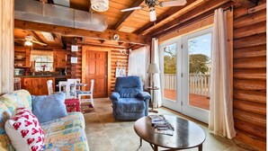 A cozy log cabin interior with a dining area, a blue armchair, a colorful sofa, and a large sliding glass door leading to a deck.