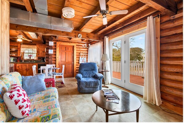 L’intérieur chaleureux d’un chalet en bois comprend un coin repas, un fauteuil bleu, un canapé coloré et une grande porte-fenêtre coulissante donnant sur une terrasse.