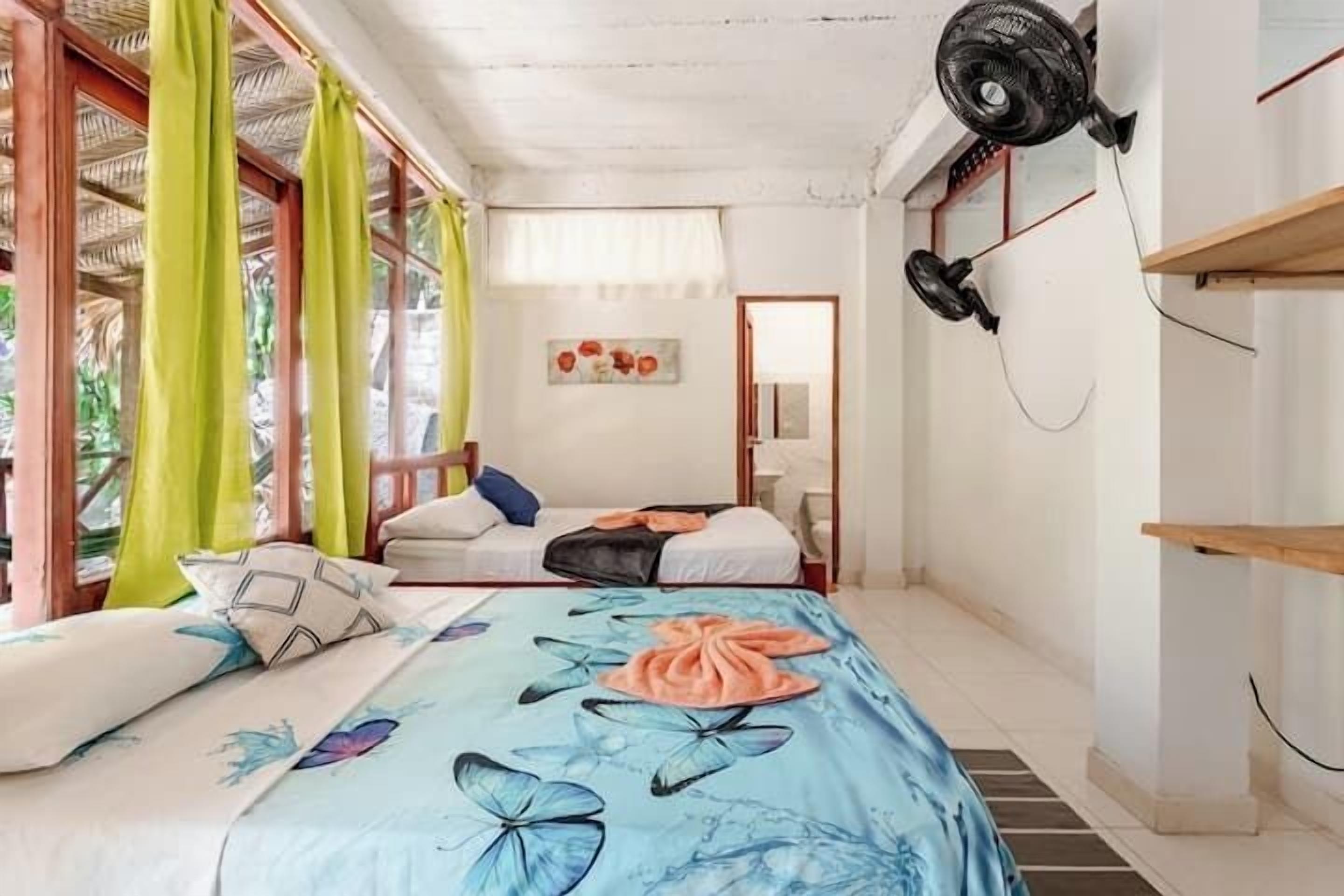 A bedroom with a bed, two fans, a window with curtains, and a door.