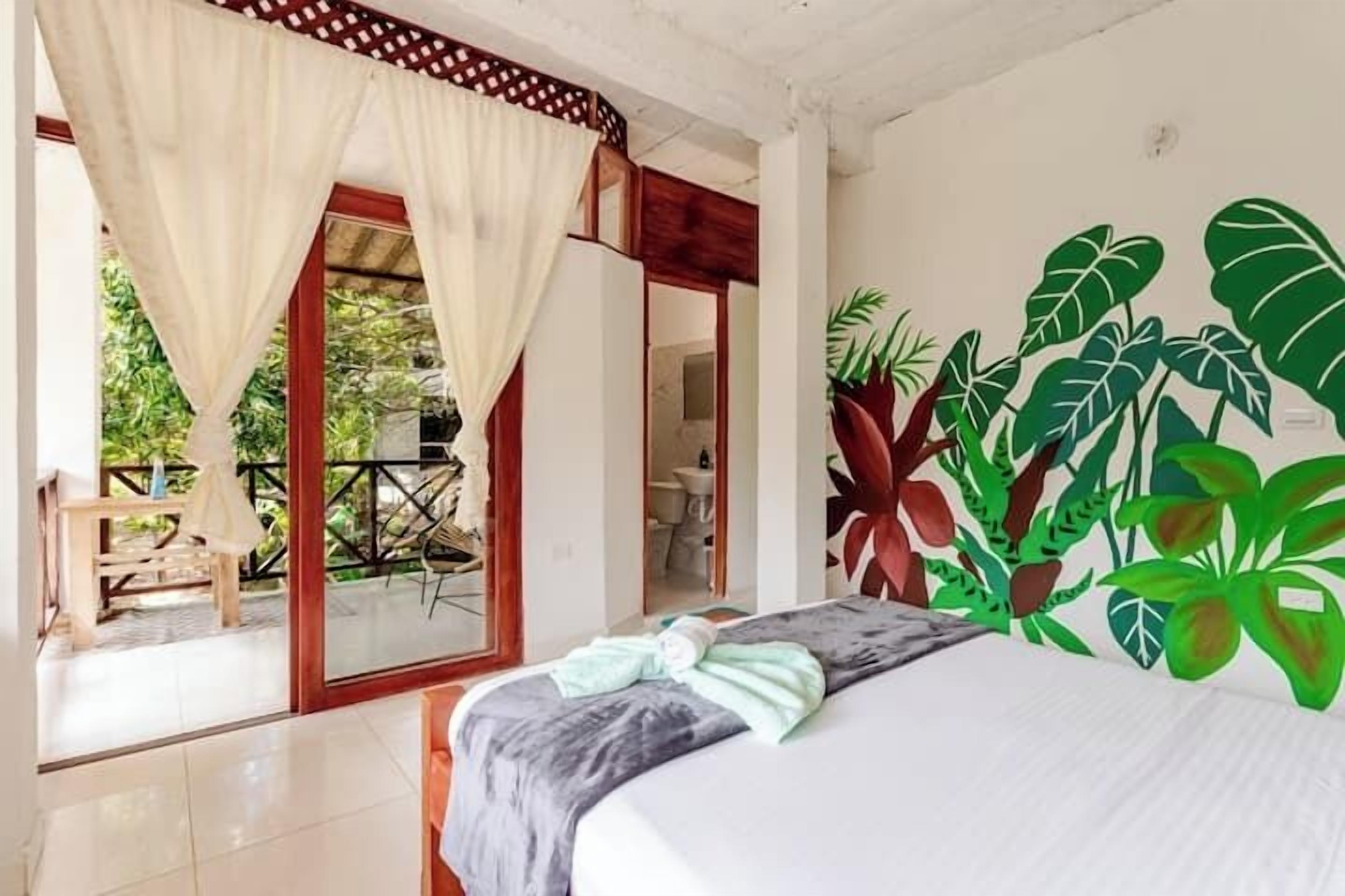 Une chambre à coucher avec un lit, des rideaux, une vue sur l’extérieur et un mur décoré d’une fresque représentant des plantes.