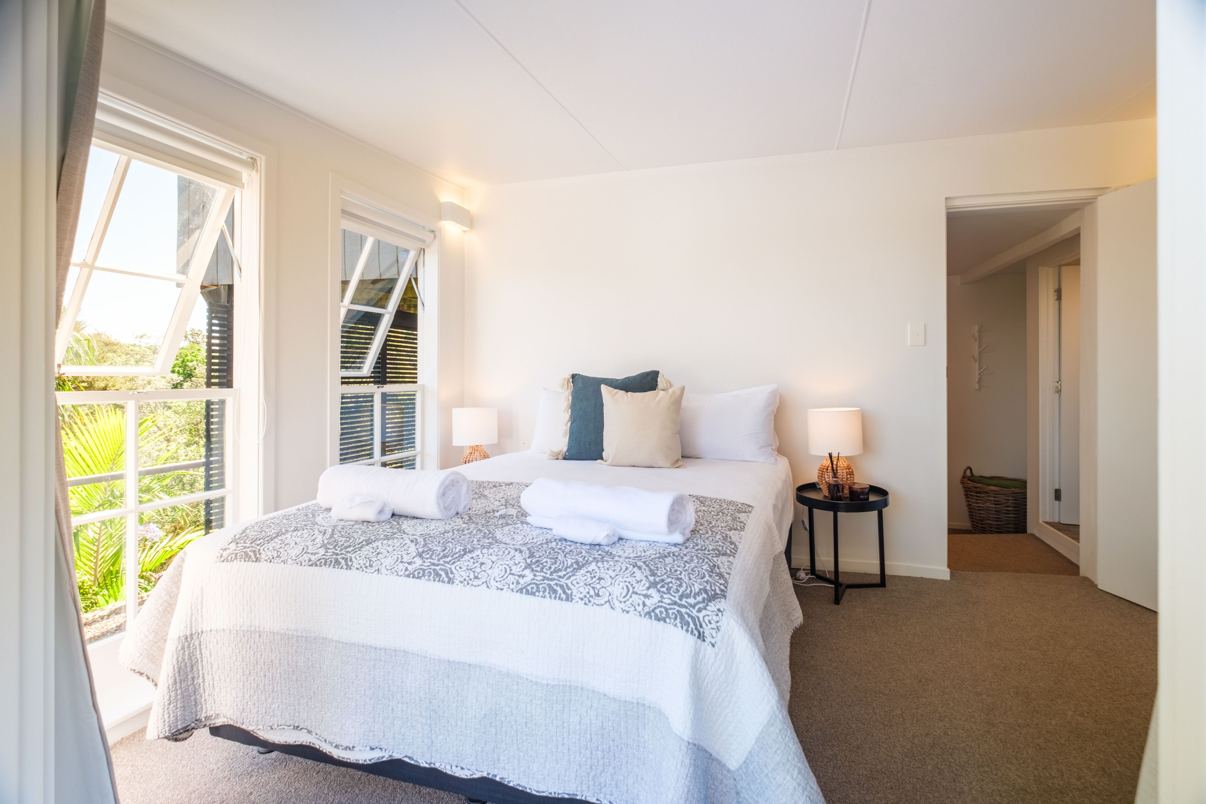 A bedroom with a large bed, two bedside tables with lamps, a window with blinds, and a view of greenery outside.