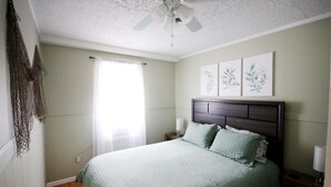 Une chambre à coucher comprenant un lit, une table de chevet, un ventilateur de plafond et des œuvres d’art accrochées au mur.