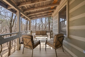 A covered porch with two chairs and a table, overlooking a forested area.