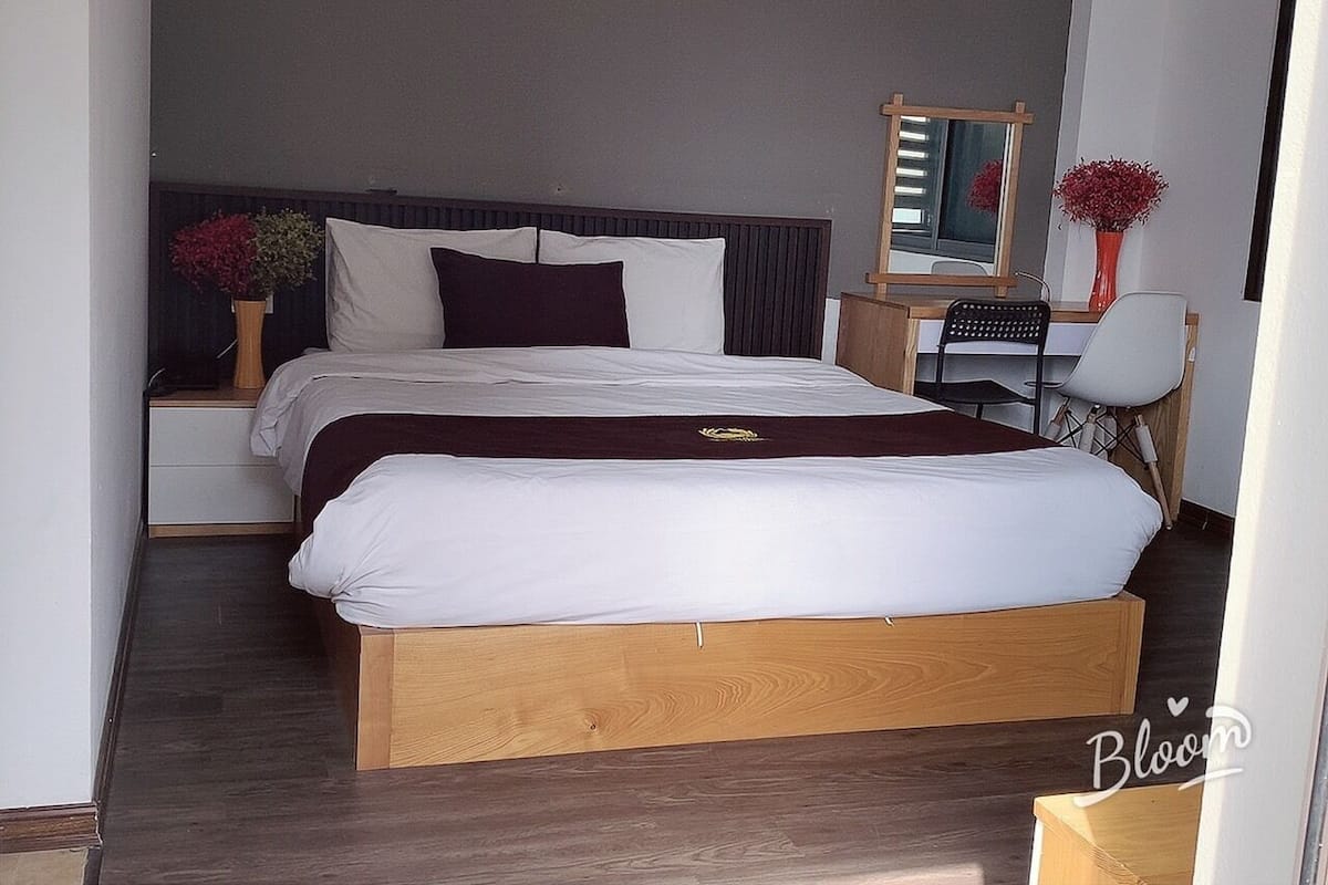 A double bed with a wooden frame, white bedding, and dark pillows. There is a nightstand with a vase of flowers and a mirror. A desk with a chair is visible in the corner.