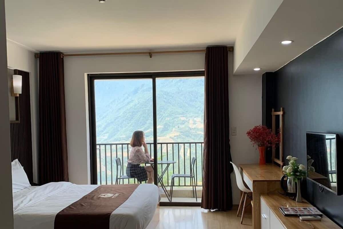 A hotel room with two beds, a desk, a TV, and a balcony with a view of the mountains.