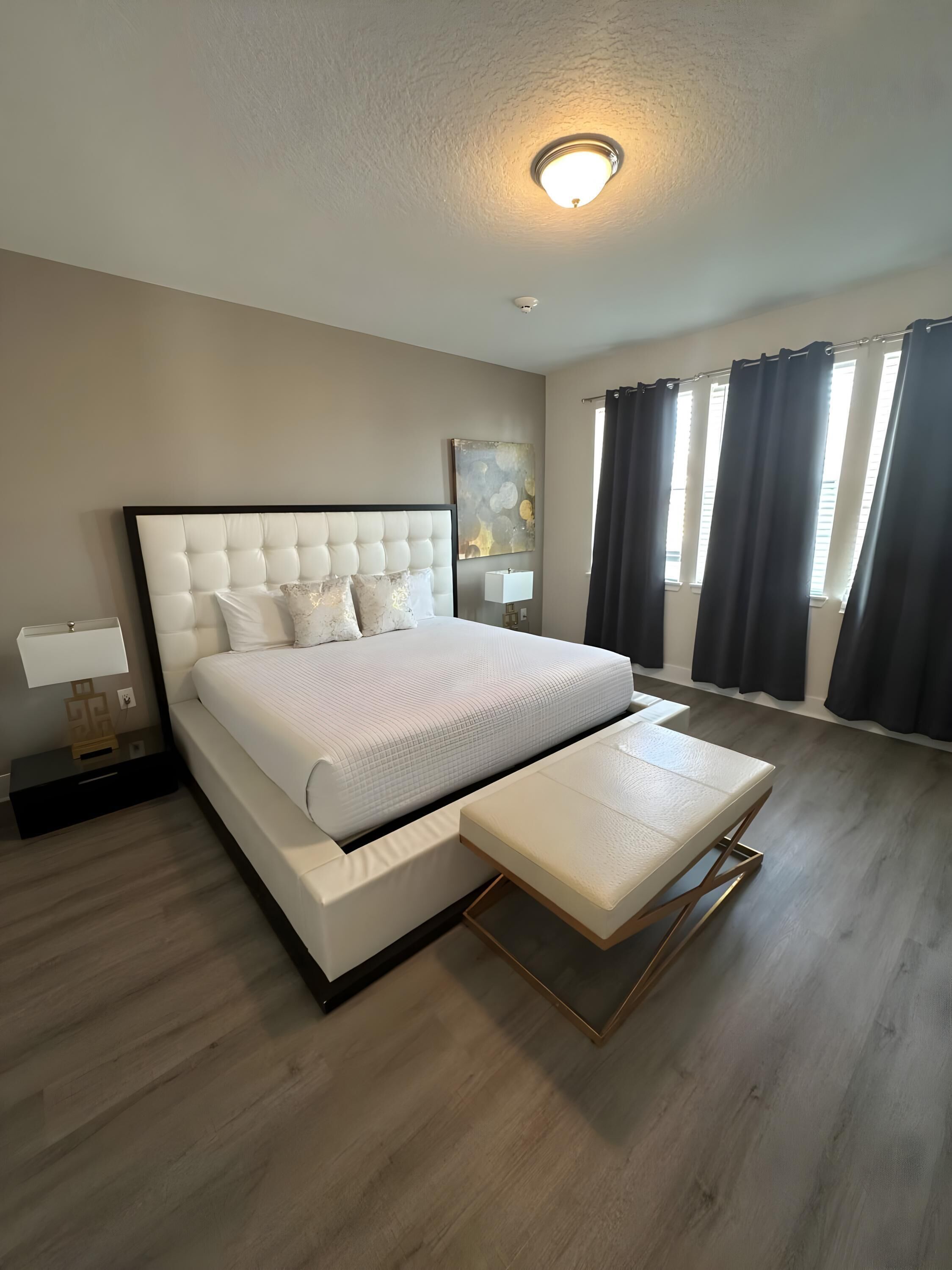 A modern bedroom with a large bed, a bedside table, a nightstand, a ceiling light, a window with curtains, and a wall-mounted artwork.