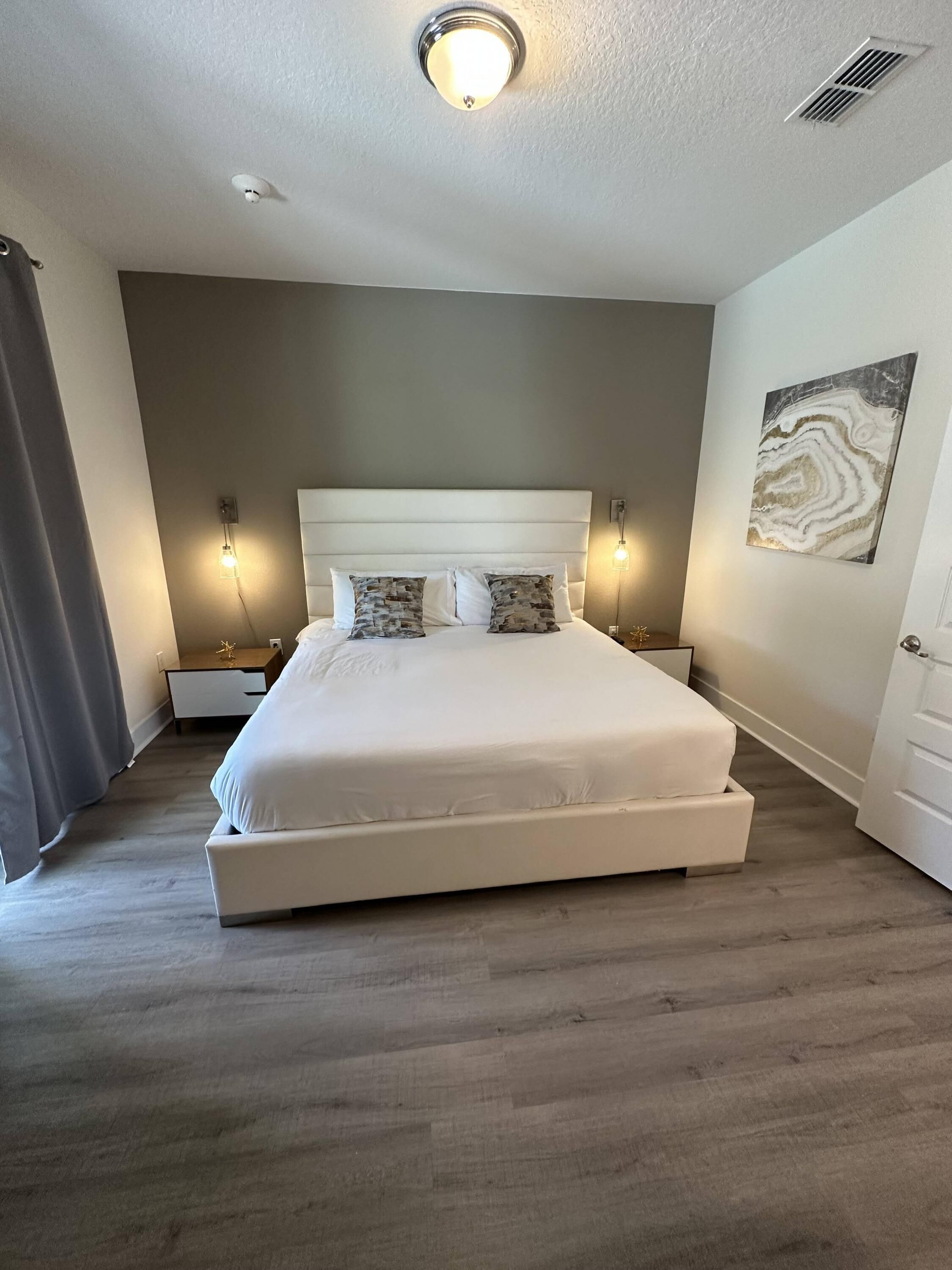 A large bed with a white headboard and two decorative pillows, a bedside table with a lamp, and a wall-mounted artwork.
