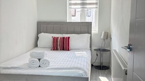 A neatly made bed with white bedding and a red and white striped pillow, a small bedside table with a lamp, and a window with blinds. - 2 Bed-Sleeps 5-Free Parking-Pet Friendly (High Wycombe)