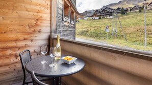 A wooden cabin with a balcony, a table with two wine glasses and a bottle, and a view of a mountain village.