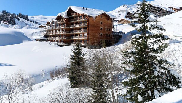 A multi-story wooden building with balconies, surrounded by snow-covered mountains and trees.