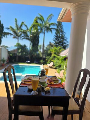 A dining table with breakfast items and a view of a swimming pool and palm trees. - Sprunger Avenue-Tulia Homes  (Moshi)