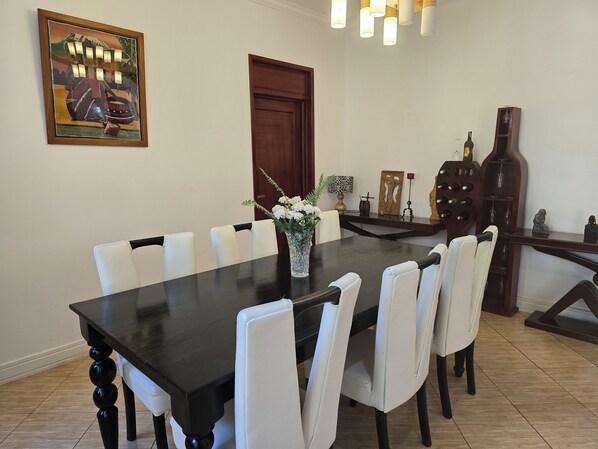 A dining room with a dark wooden table, white chairs, a vase with flowers, and a painting on the wall. - Sprunger Avenue-Tulia Homes  (Moshi)