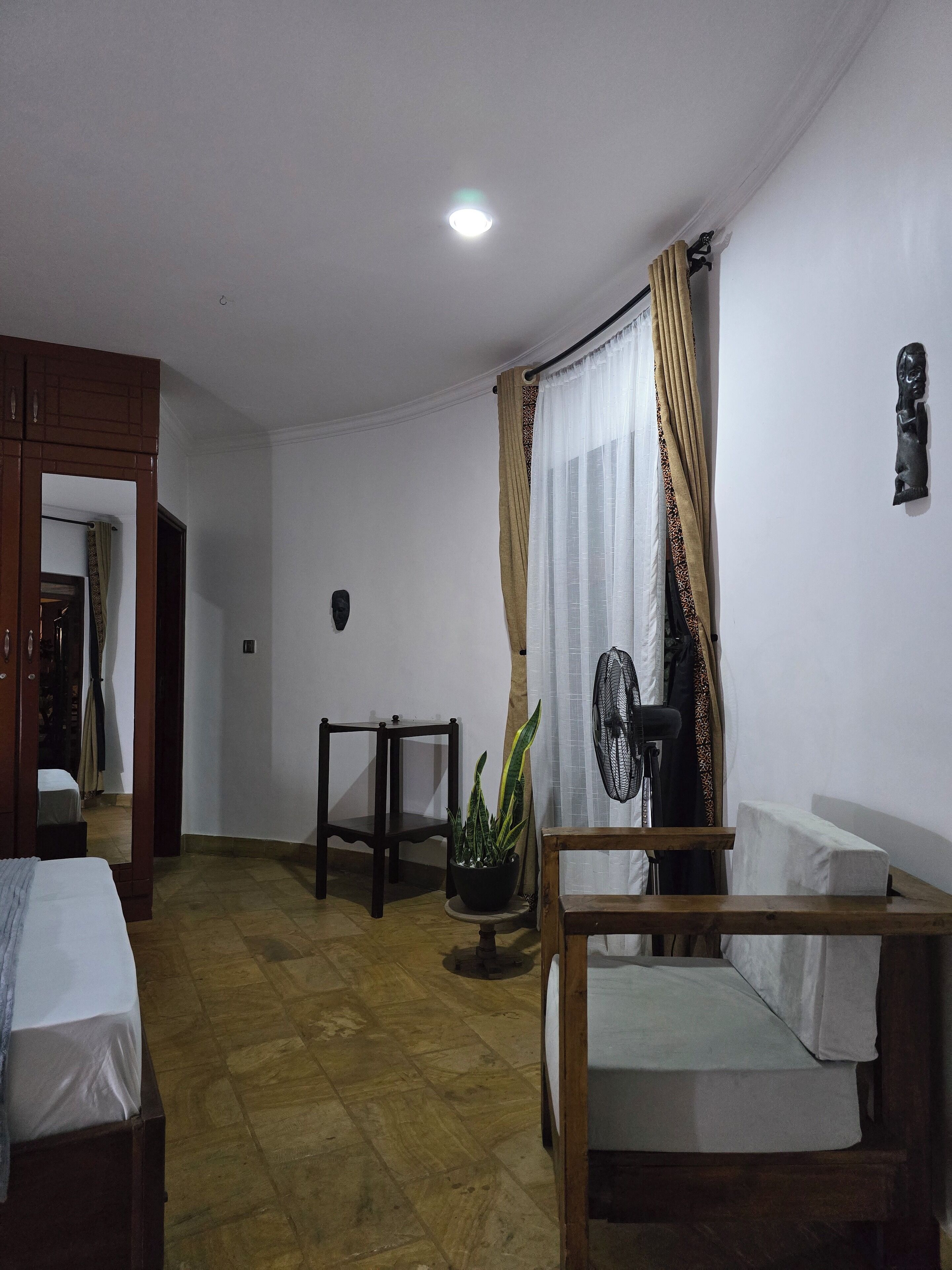 A room with a bed, a small table, a potted plant, a fan, and a curtain.