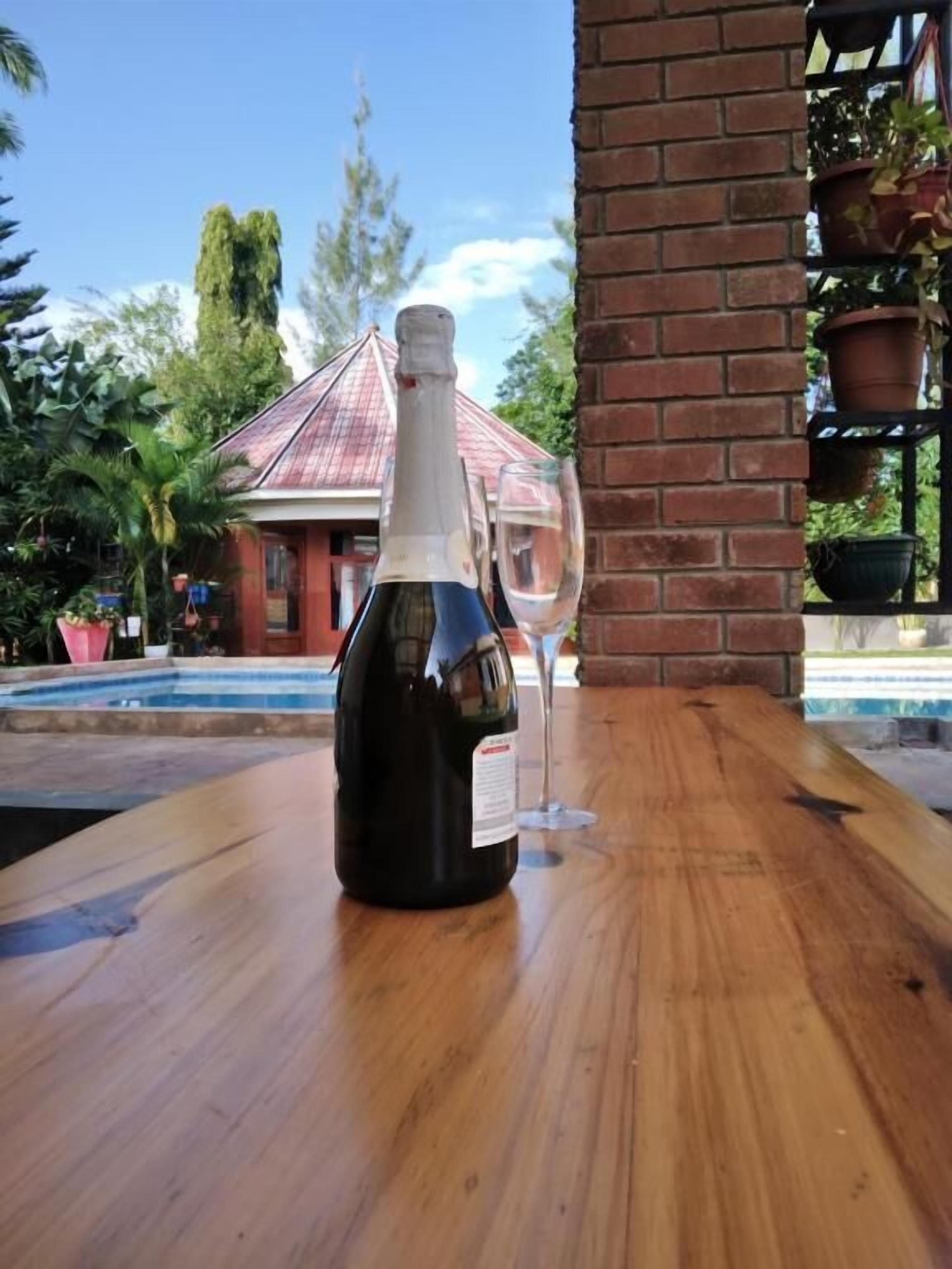 A bottle of wine on a wooden table with a glass beside it, set against a backdrop of a building with a red roof and a pool.