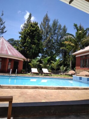 A swimming pool with lounge chairs, surrounded by trees and a building with a red roof. - Sprunger Avenue-Tulia Homes  (Moshi)