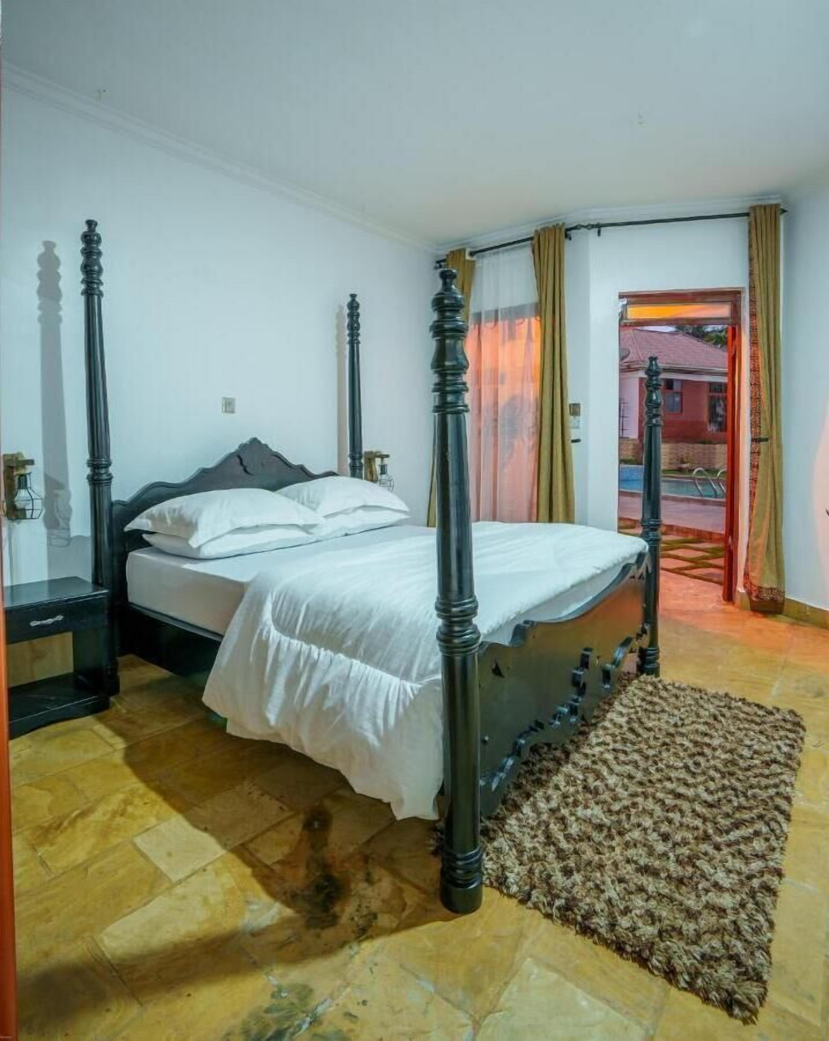 A bedroom with a four-poster bed, a wooden floor, and a view of the outside through a doorway.