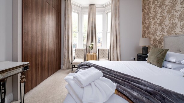 A hotel room with a large bed, a wooden wardrobe, a desk, and a chair. - GuestReady - Fabulous stay in Fulham (London)