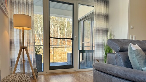 Een moderne woonkamer met een grijze bank, een houten vloer, een lamp en uitzicht door een glazen deur op een buitenruimte.