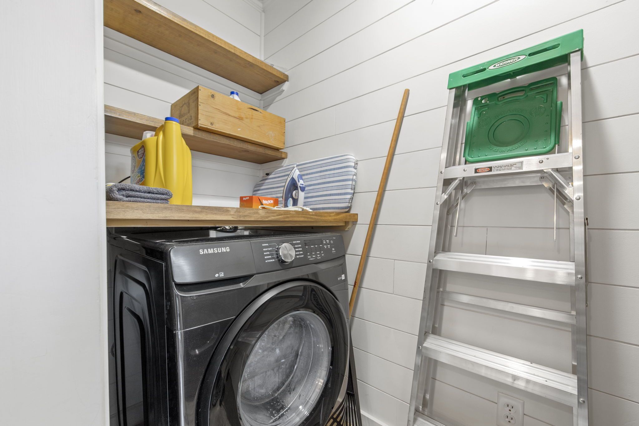 A laundry area with a Samsung washing machine, wooden shelves, and a green ladder.