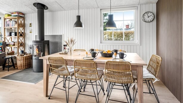 A cozy dining area with a large wooden table, wicker chairs, and a black pendant light. A black stove is visible in the background, and there is a bookshelf filled with books and decorative items.