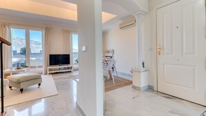 Amplia sala de estar con televisor de pantalla plana, comedor con mesa y sillas blancas, y balcón con vista a las montañas.
