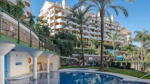 Una piscina con superficie de agua azul cristalina, rodeada de palmeras y un edificio de varios pisos con balcones.
