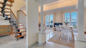 Amplia sala de estar con escalera, comedor con sillas blancas y vista al exterior.