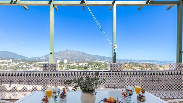 Una mesa dispuesta con fruta, vasos de jugo y un florero con plantas, con vista a una montaña y un pueblo al fondo.